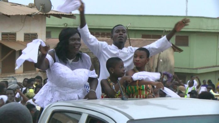 Father Stephen in a plain white cassock, his mother in a white kaba with black stripes and a bright headgear.